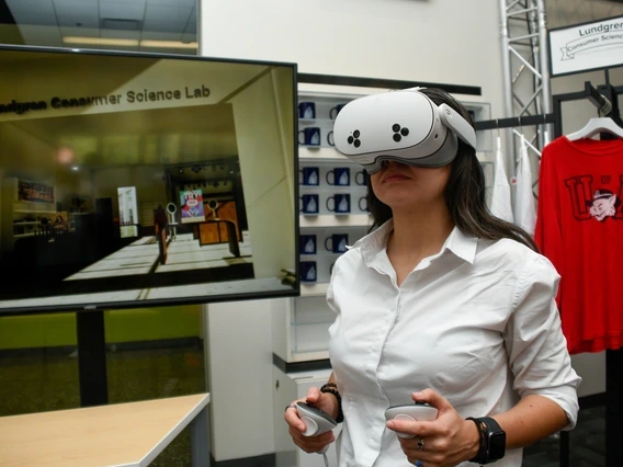A woman wearing a white button-down shirt uses a virtual reality headset and handheld controllers inside the Lundgren Consumer Science Lab. A large screen behind her displays a virtual retail space, and shelves of mugs and a red "U of A" shirt are visible in the background.