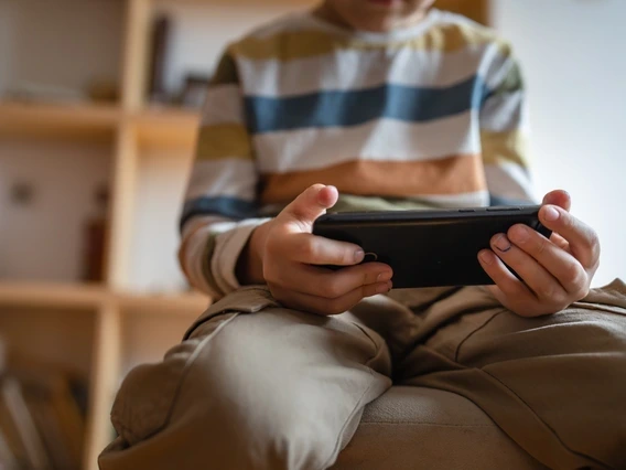 A young child wearing a striped long-sleeve shirt and khaki pants sits indoors, intently holding and looking at a smartphone. The focus is on the child’s hands and device, with a blurred background of shelves and books