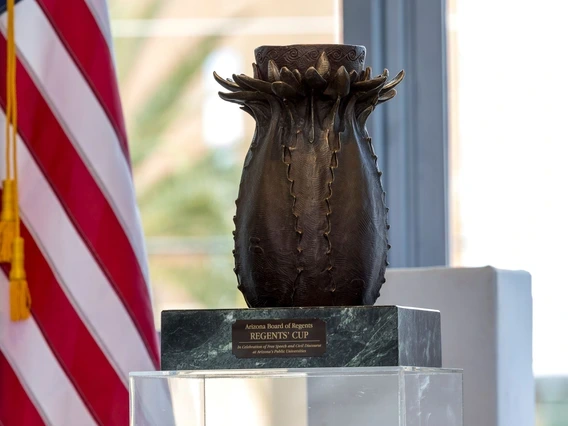 A bronze trophy shaped like a stylized cactus sits on a dark green marble base, with a plaque that reads: "Arizona Board of Regents REGENTS' CUP – In Celebration of Free Speech and Civil Discourse at Arizona’s Public Universities." The trophy is displayed in front of a blurred American flag and a window.
