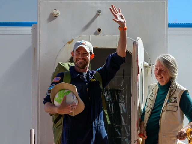 A person opens the door to a sealed habitat as a second person exits, waving and smiling.