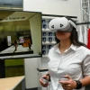 A woman wearing a white button-down shirt uses a virtual reality headset and handheld controllers inside the Lundgren Consumer Science Lab. A large screen behind her displays a virtual retail space, and shelves of mugs and a red "U of A" shirt are visible in the background.